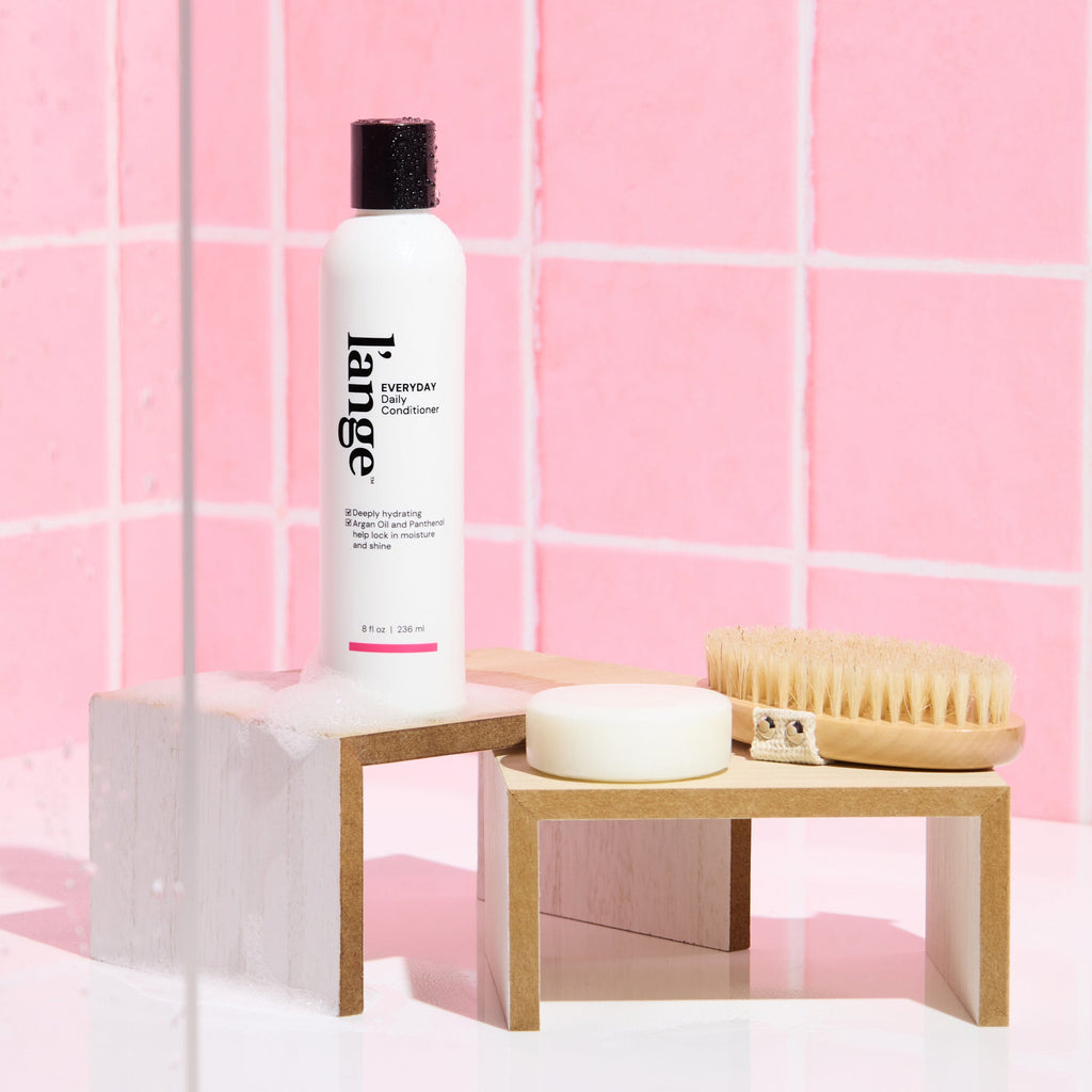 L’ange Everyday Conditioner on a wooden stand against a pink tile background with soap suds