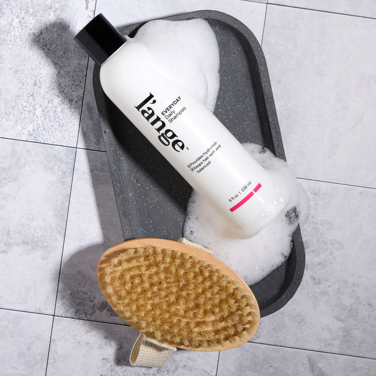 Overhead view of L’ange Shampoo with a body brush and clips on a grey tile surface