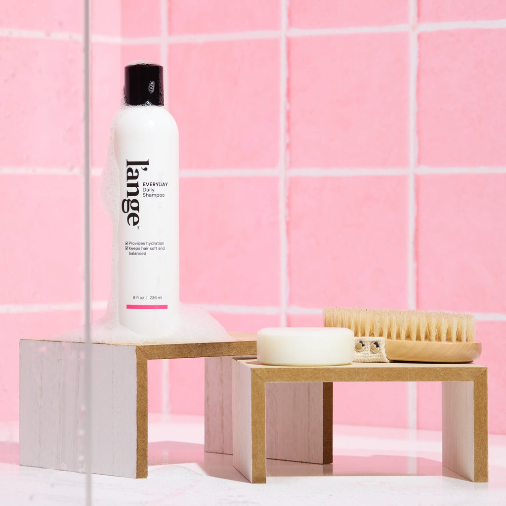 L’ange Everyday Shampoo on a wooden stand against a pink tile background with soap suds