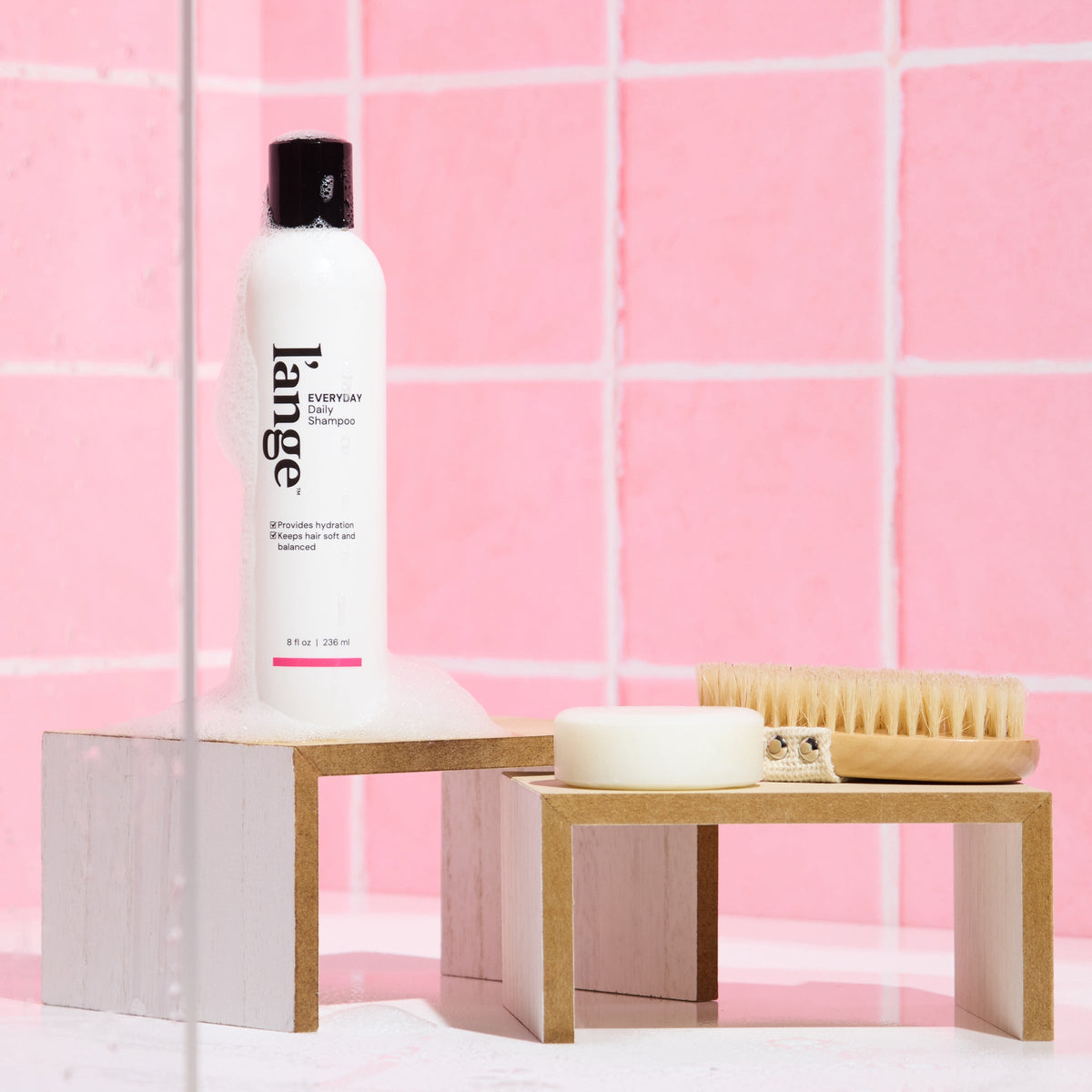 L’ange Everyday Shampoo on a wooden stand against a pink tile background with soap suds