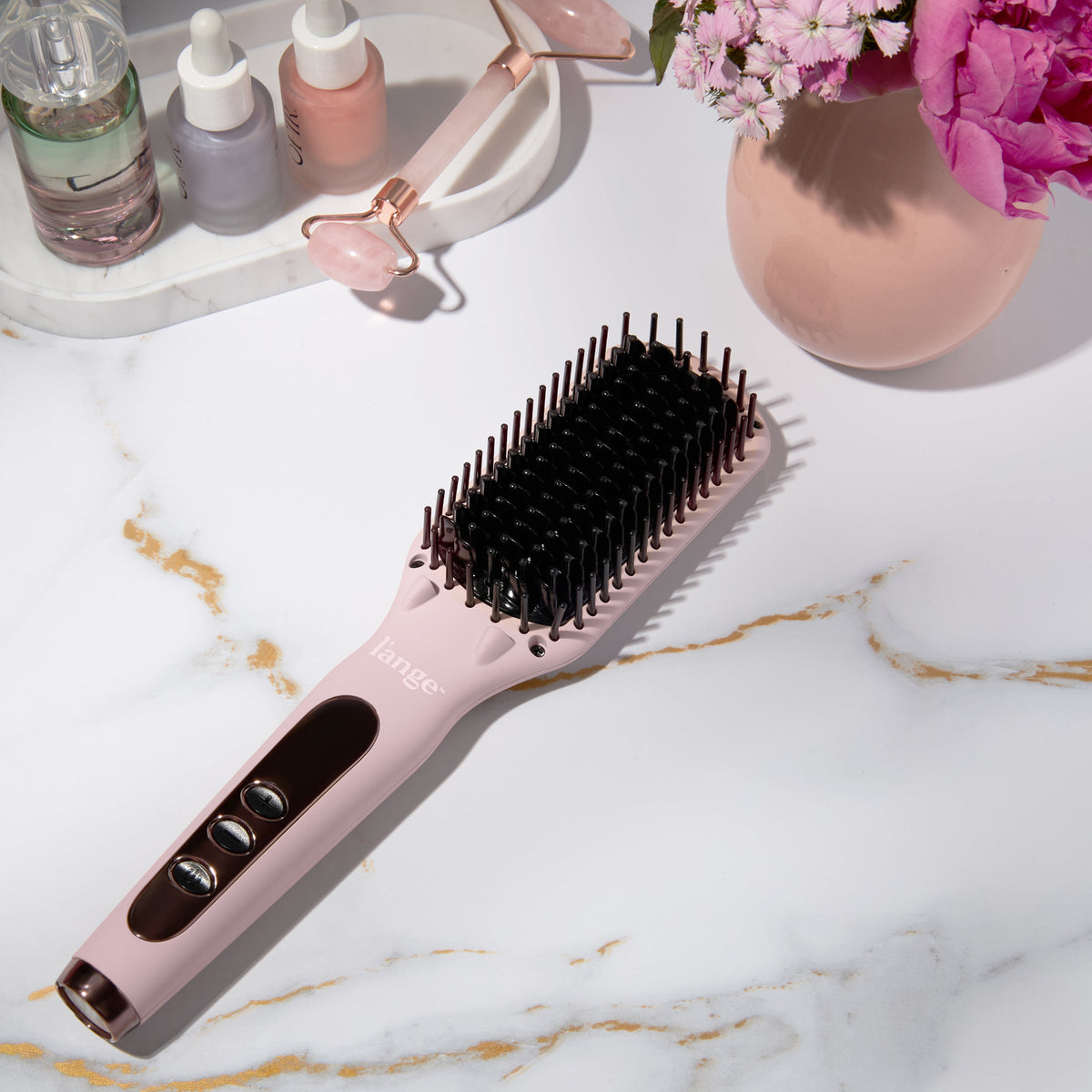 Blush heated straightening brush on a marble counter beside beauty products and flowers.
