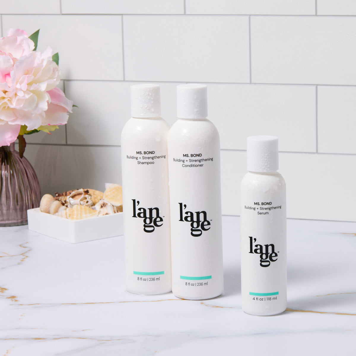 L'ange MS. Bond haircare collection displayed includes building and strengthening shampoo, conditioner, and serum in sleek white bottles on a marble countertop, accented with a vase of pink flowers and seashells.