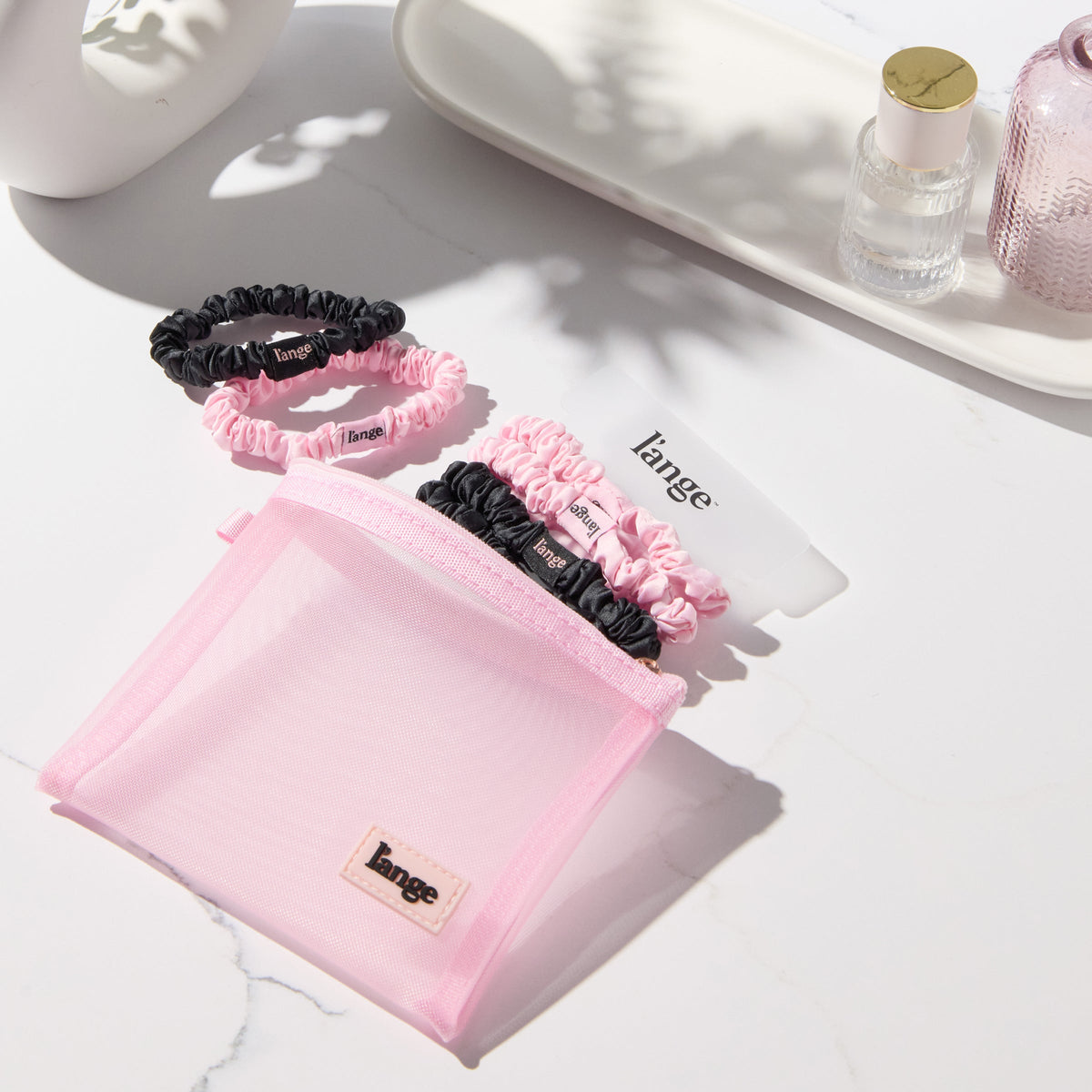 blush and black L’ange satin scrunchies on a vanity beside a small pink zip pouch