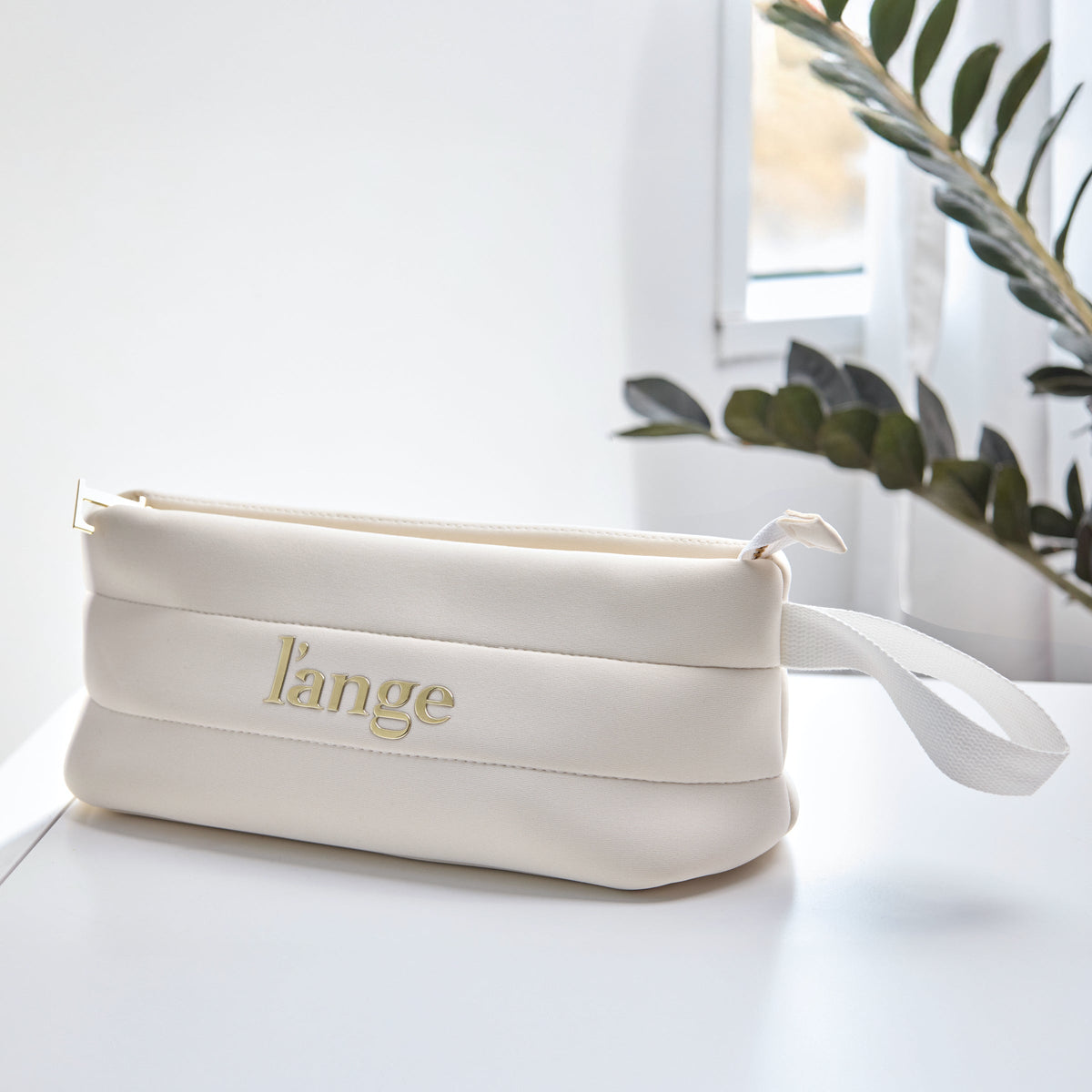 Whhite L’ange zippered cosmetic bag with gold logo and wrist strap on a white surface.